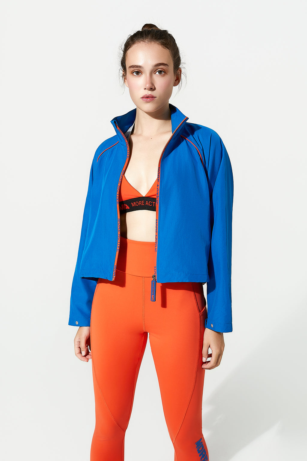 Woman wearing a blue jacket and orange athletic Lila gym leggings sculpt, support, and move with you. High-rise fit, phone pocket, lightweight comfort. Train hard. Shop now. on a white background