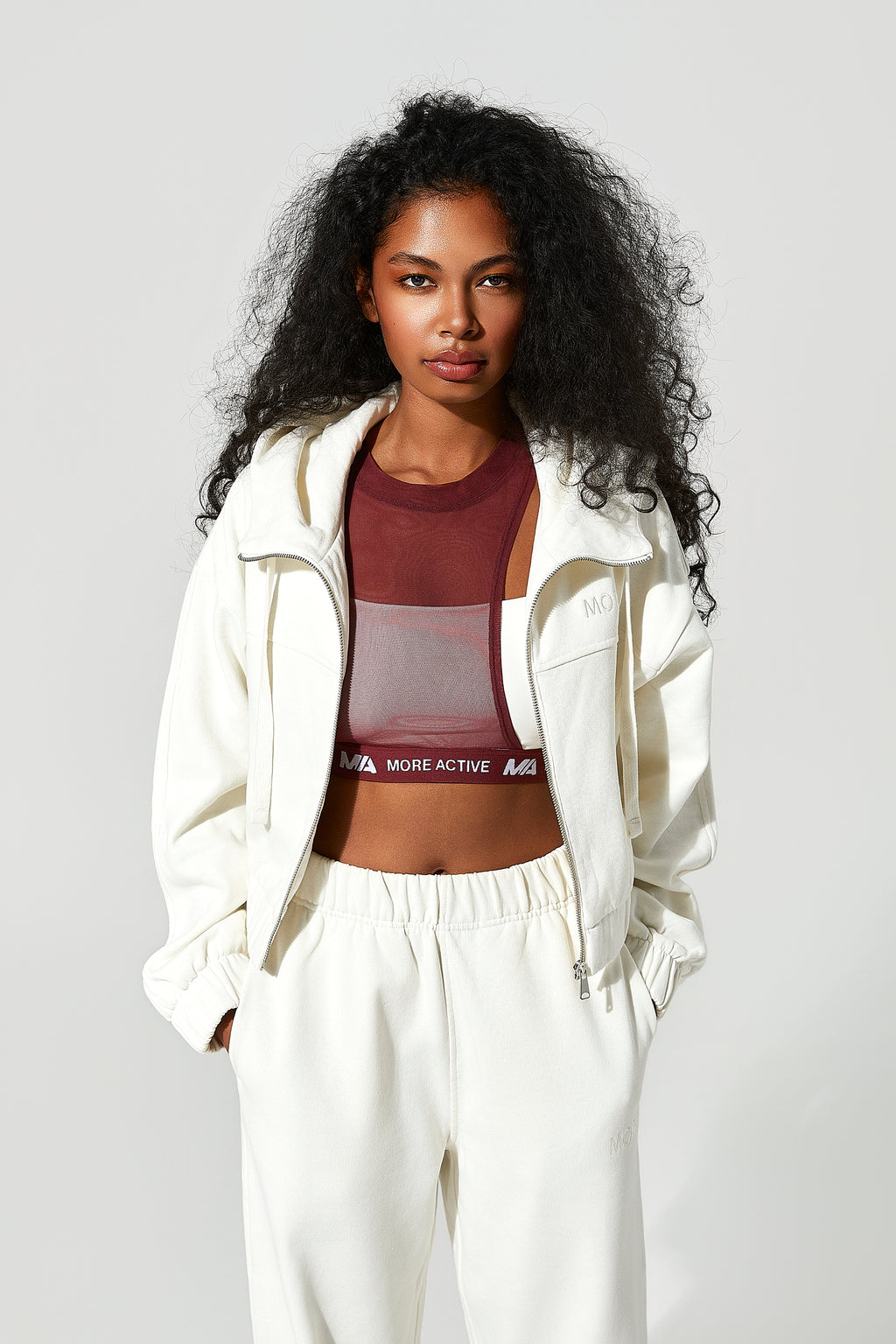 Woman wearing a white tracksuit with a maroon sports bra featuring 'More Active' branding on a light gray background.
