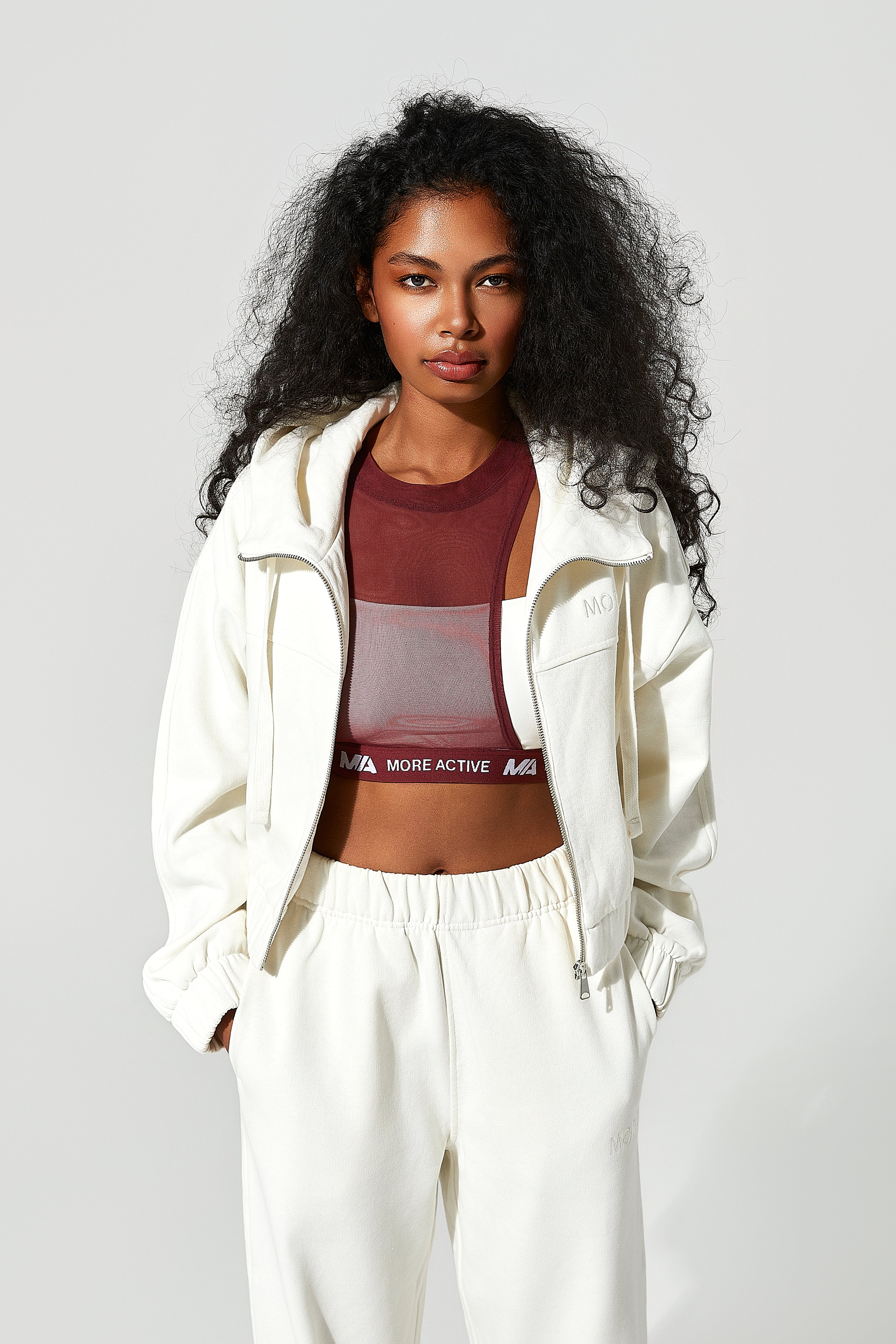 Woman wearing a white tracksuit with a maroon sports bra featuring 'More Active' branding on a light gray background.