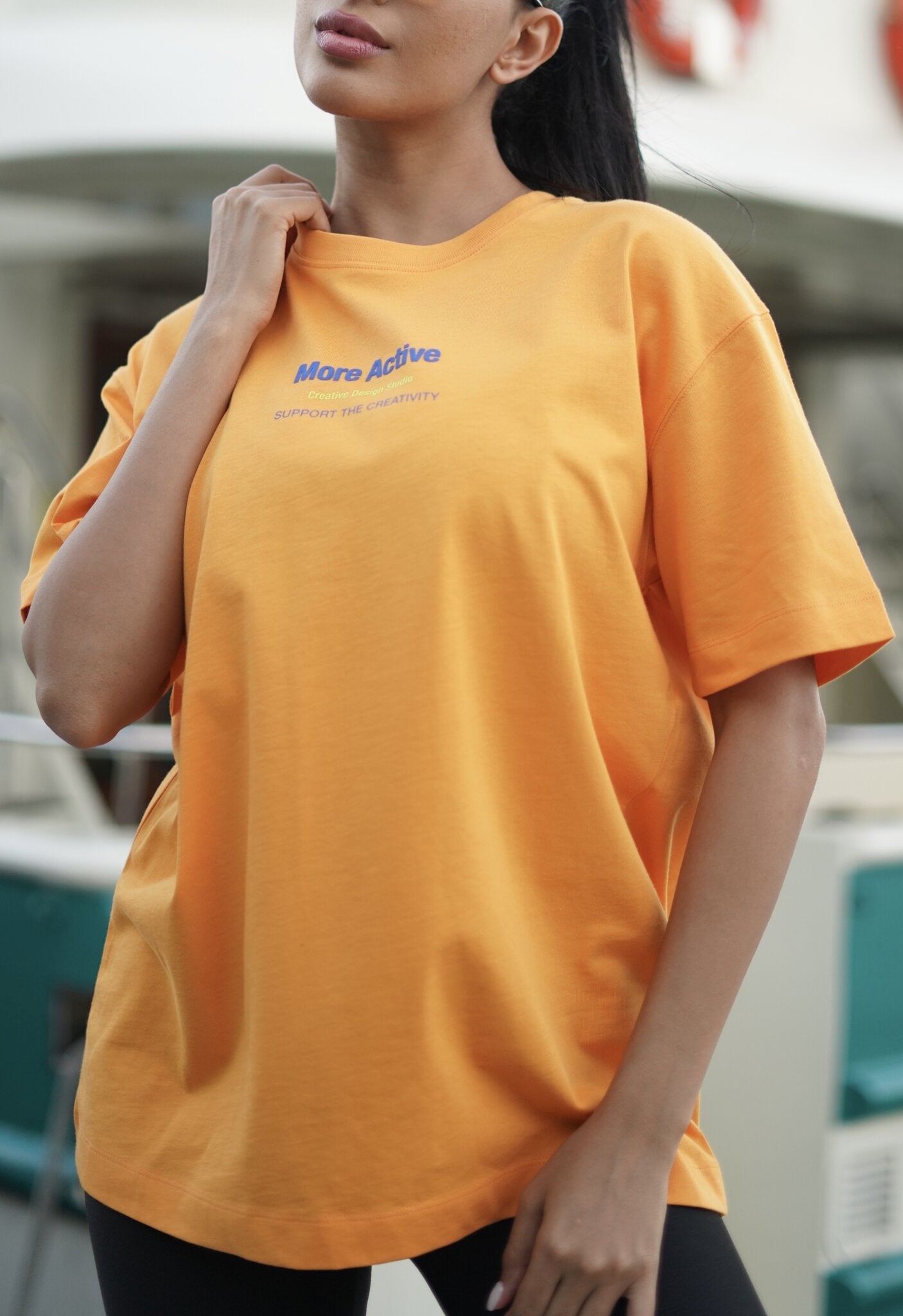 A woman wearing an oversized yellow t-shirt with blue text that reads 'More Active' and 'empowering the community' on the chest, set against a blurred outdoor background.