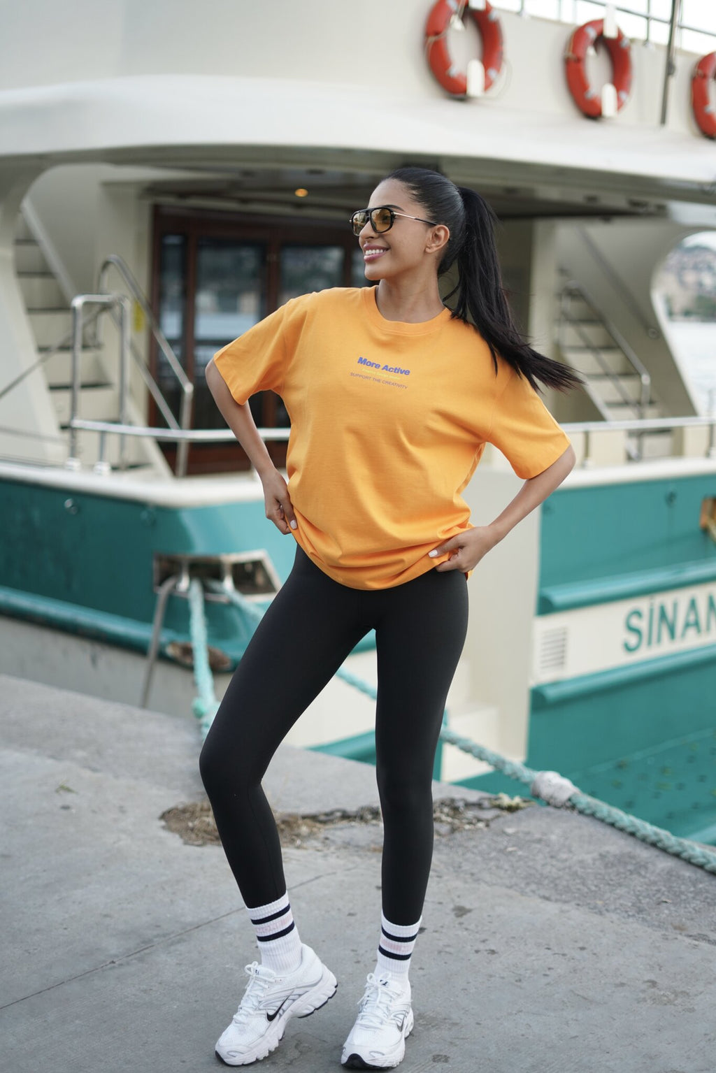 A woman posing on a dock in front of a teal and white boat, wearing a bright yellow t-shirt with a small blue graphic, black leggings, white socks, and white sneakers.