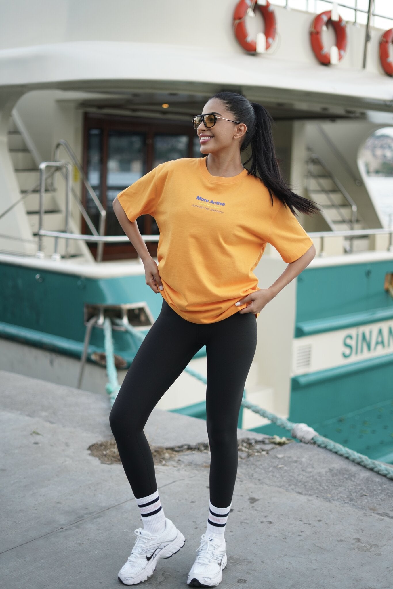 A woman posing on a dock in front of a teal and white boat, wearing a bright yellow t-shirt with a small blue graphic, black leggings, white socks, and white sneakers.