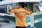 A woman wearing a bright orange t-shirt with blue and yellow text on the back, posing in front of a boat dock.