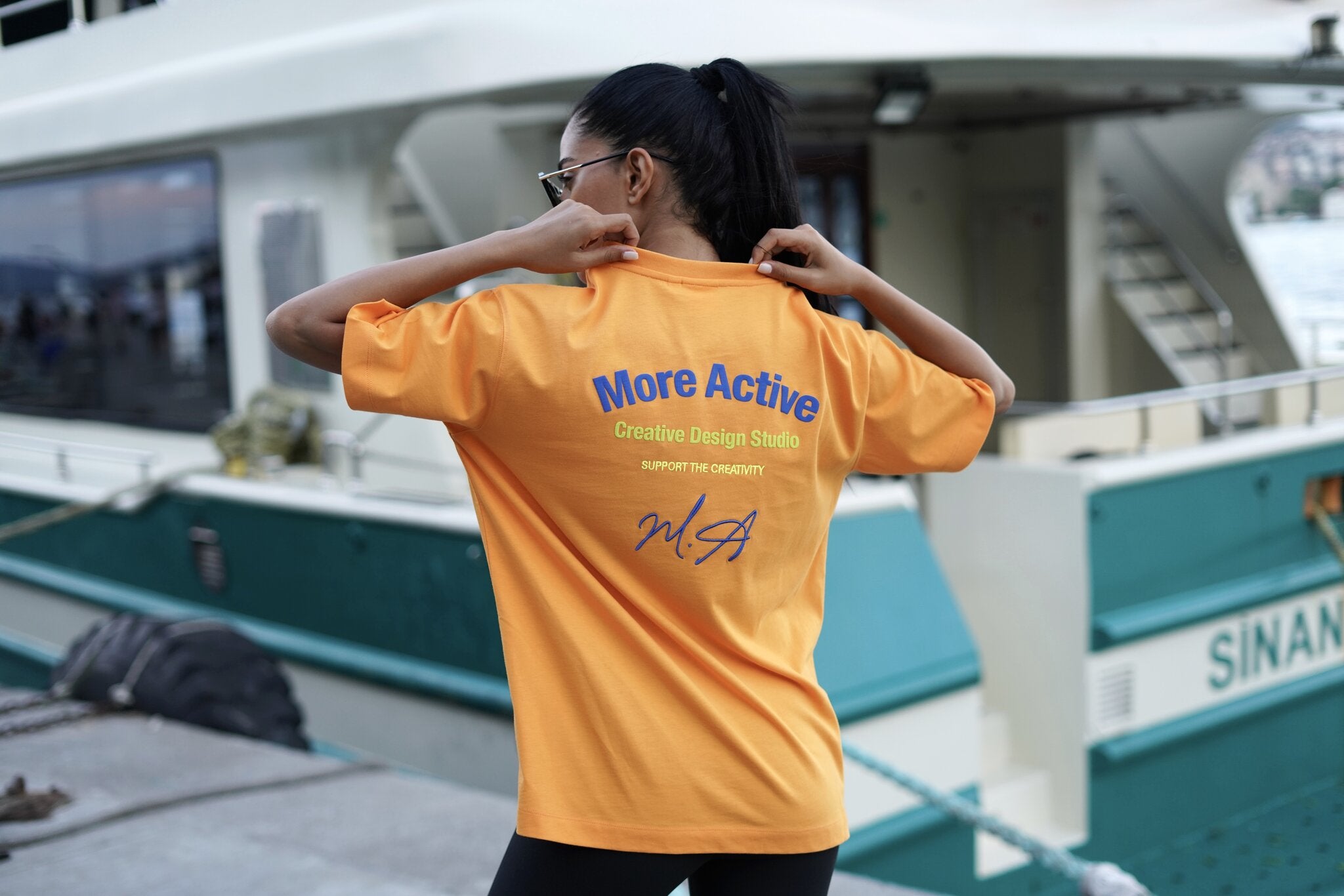 A woman wearing a bright orange t-shirt with blue and yellow text on the back, posing in front of a boat dock.