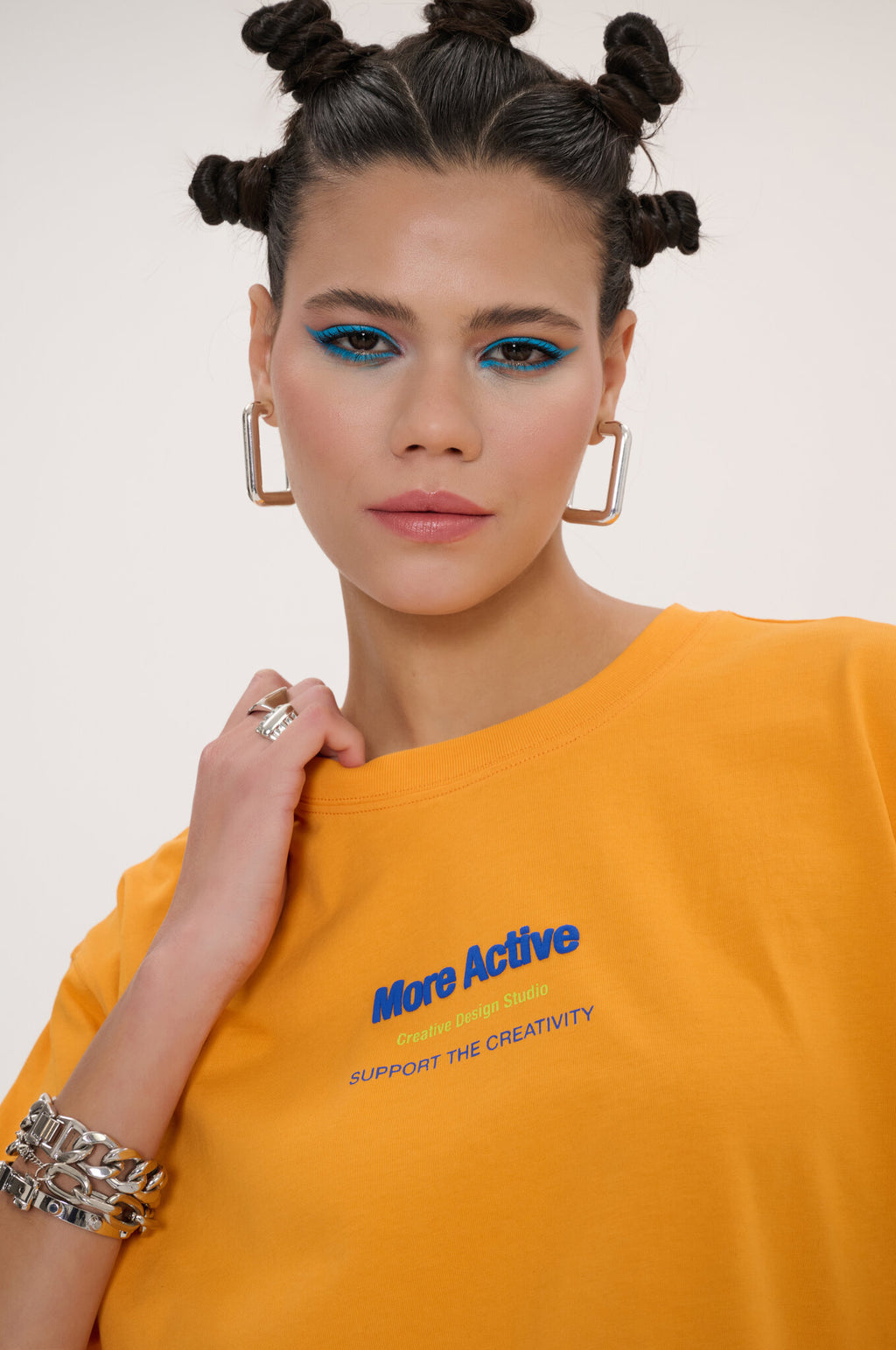 A close-up of a model wearing a bright yellow t-shirt with blue and pink text that reads 'More Active' and 'SUPPORT THE CREATIVITY'. The model has her hair in two buns and is wearing gold hoop earrings and bracelets against a plain light background.