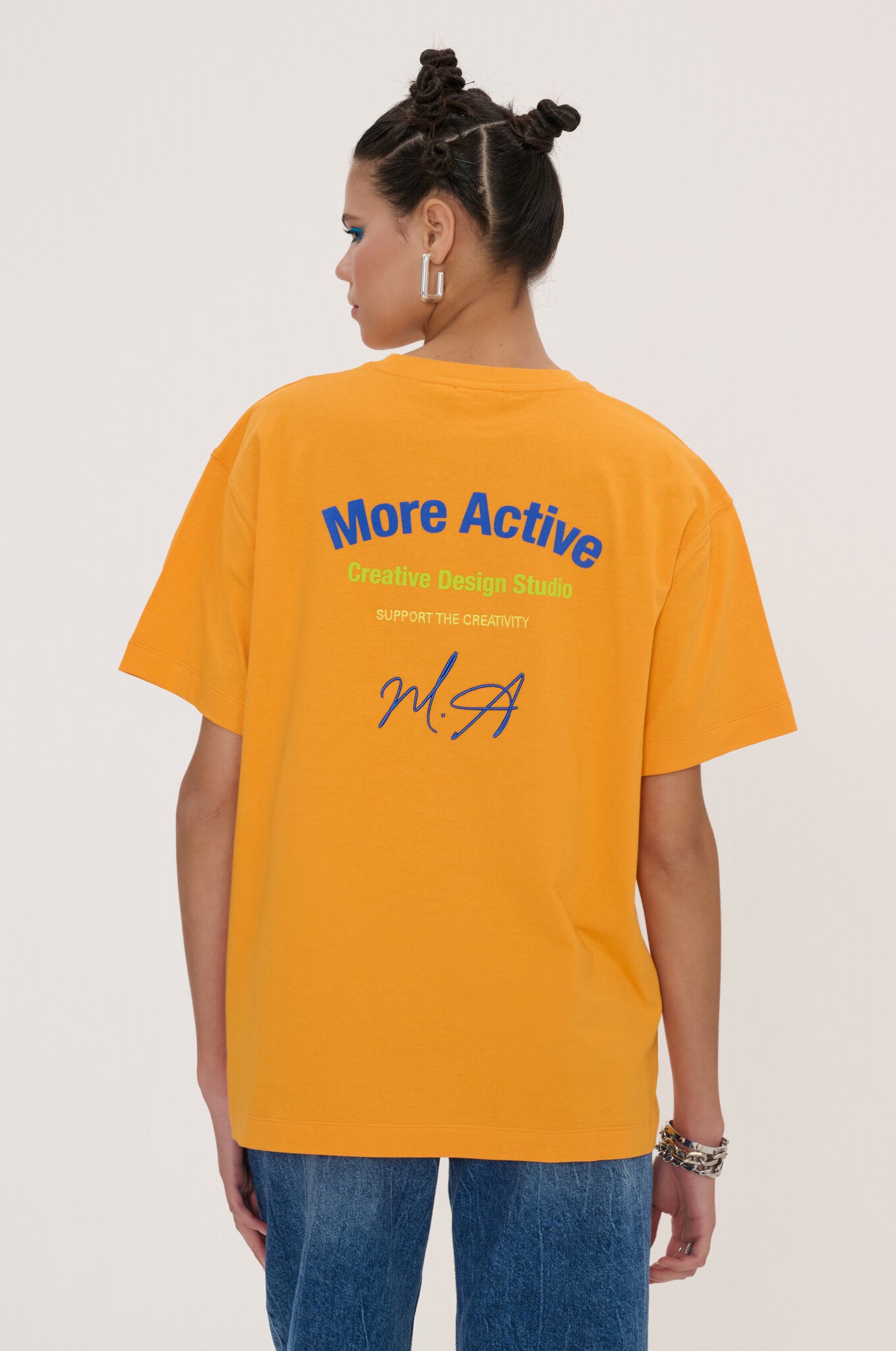 A woman shown from the back wearing an oversized bright yellow t-shirt with blue and green text on the back reading 'More Active Creative Design Studio' and a blue signature graphic. She is also wearing blue jeans, set against a plain light beige studio background.
