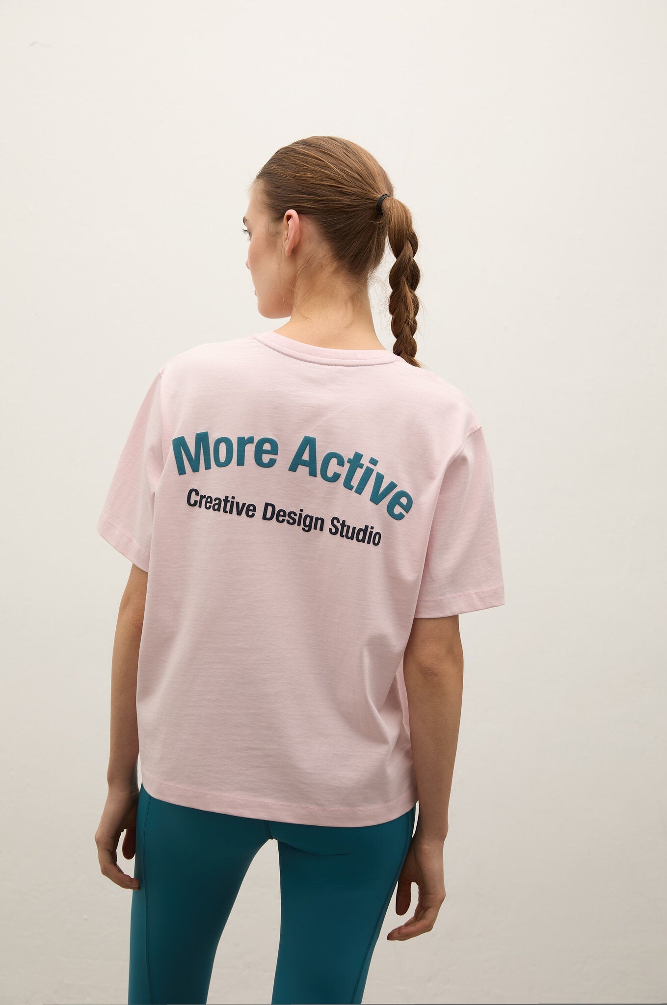 A woman viewed from behind wearing a light pink short-sleeve T-shirt with the text 'More Active Creative Design Studio' printed in teal and black on the back, paired with teal leggings, against a plain off-white studio background.