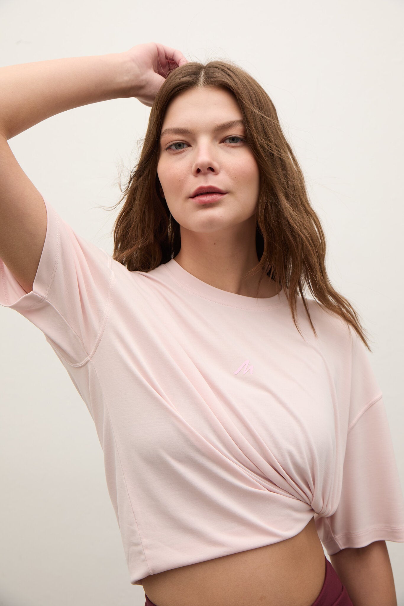 A woman wearing a light pink short-sleeve crop top with a twisted front detail, posed against a plain off-white background.