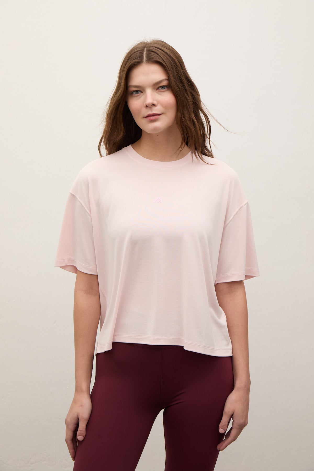 A woman modeling a light pink short-sleeve crop top with a relaxed fit, paired with burgundy leggings, against a plain off-white background.