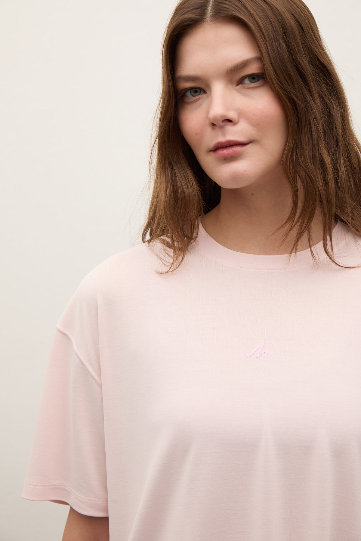 A woman wearing a pale pink oversized crewneck t-shirt with a small embroidered logo on the left chest, set against a plain light background.