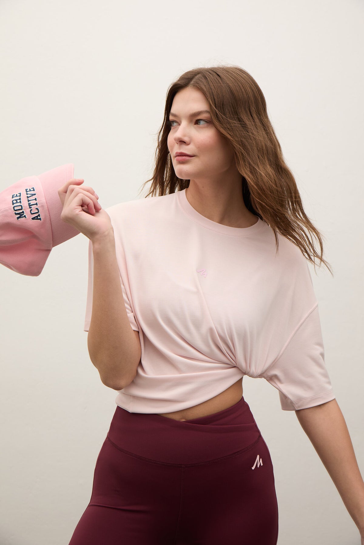 A woman modeling a light pink short-sleeve crop top with a twisted front detail, paired with burgundy leggings. She is holding a matching pink sleeve in her hand against a plain off-white studio background.