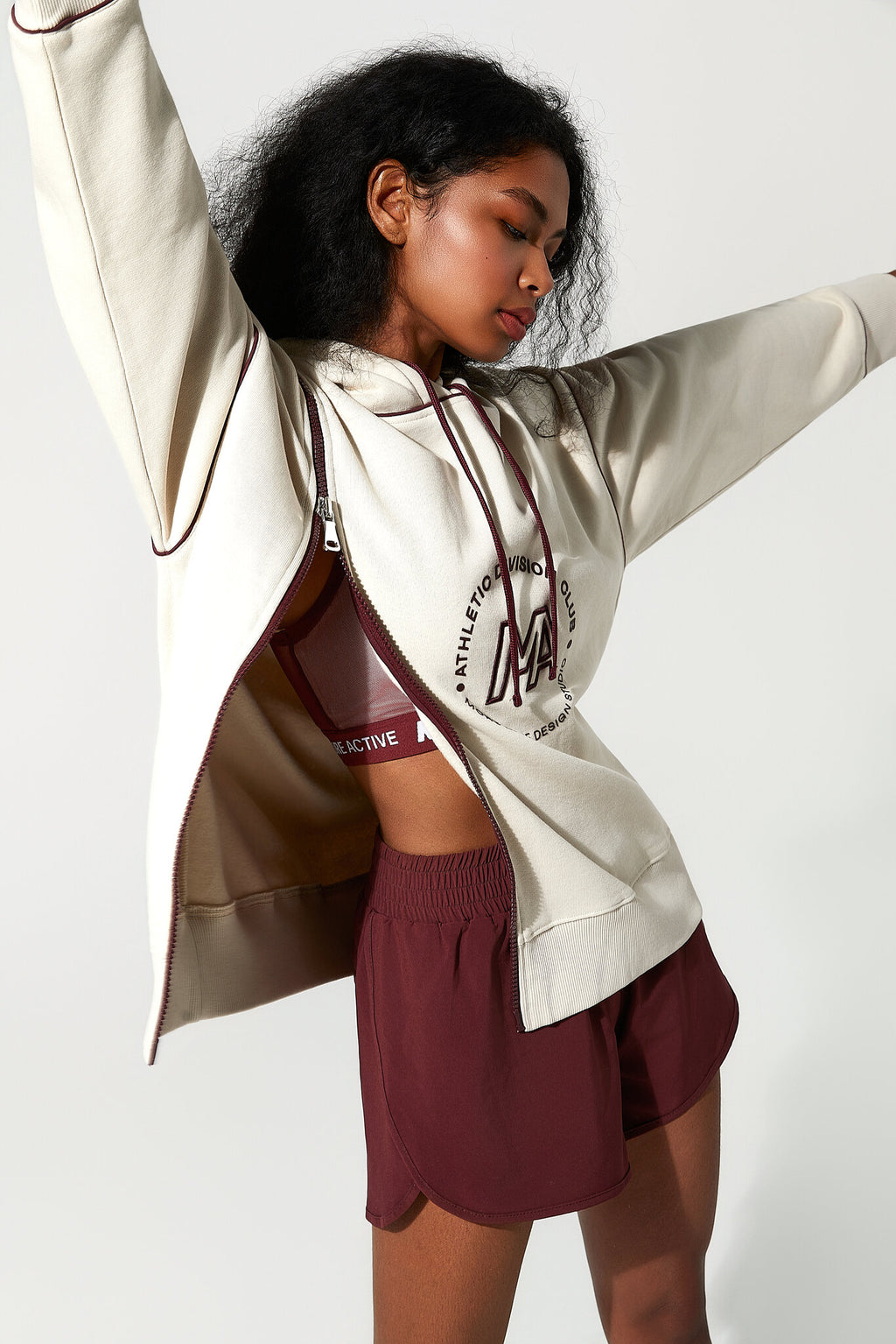 A woman modeling a cream zip-up hoodie with a burgundy circular graphic on the front, worn open over a matching burgundy sports bra and burgundy athletic shorts, against a plain light gray studio background.