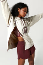 A woman modeling a cream zip-up hoodie with a burgundy circular graphic on the front, worn open over a matching burgundy sports bra and burgundy athletic shorts, against a plain light gray studio background.