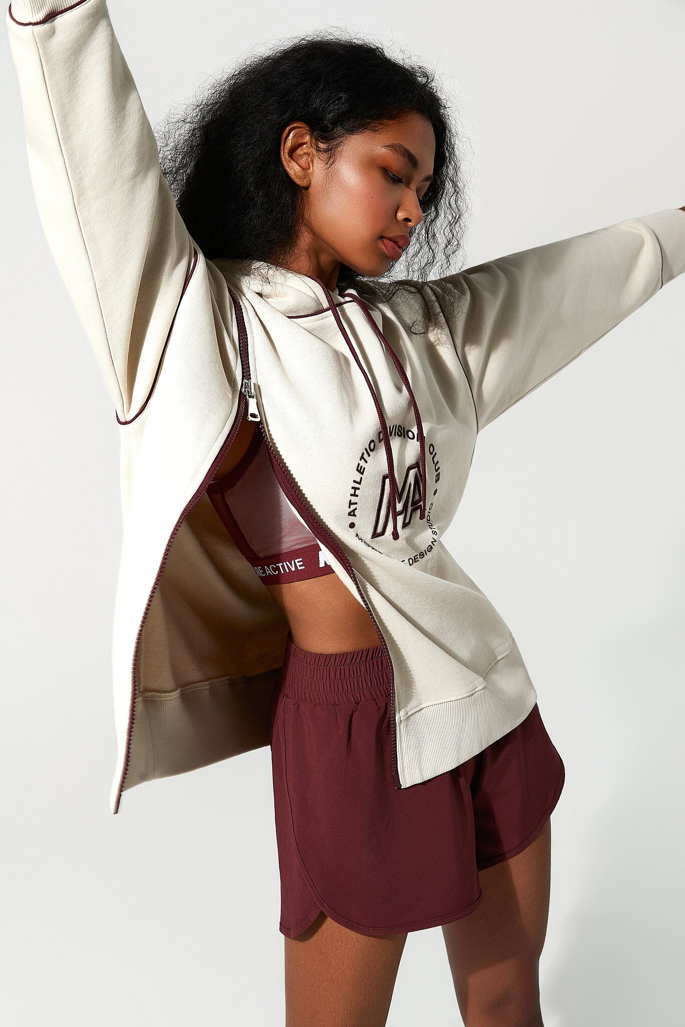 A woman modeling a cream zip-up hoodie with a burgundy circular graphic on the front, worn open over a matching burgundy sports bra and burgundy athletic shorts, against a plain light gray studio background.