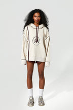 A model stands against a light gray studio background wearing an oversized cream hoodie with dark brown drawstrings and a circular chest graphic, paired with dark shorts, white socks, and gray sneakers.