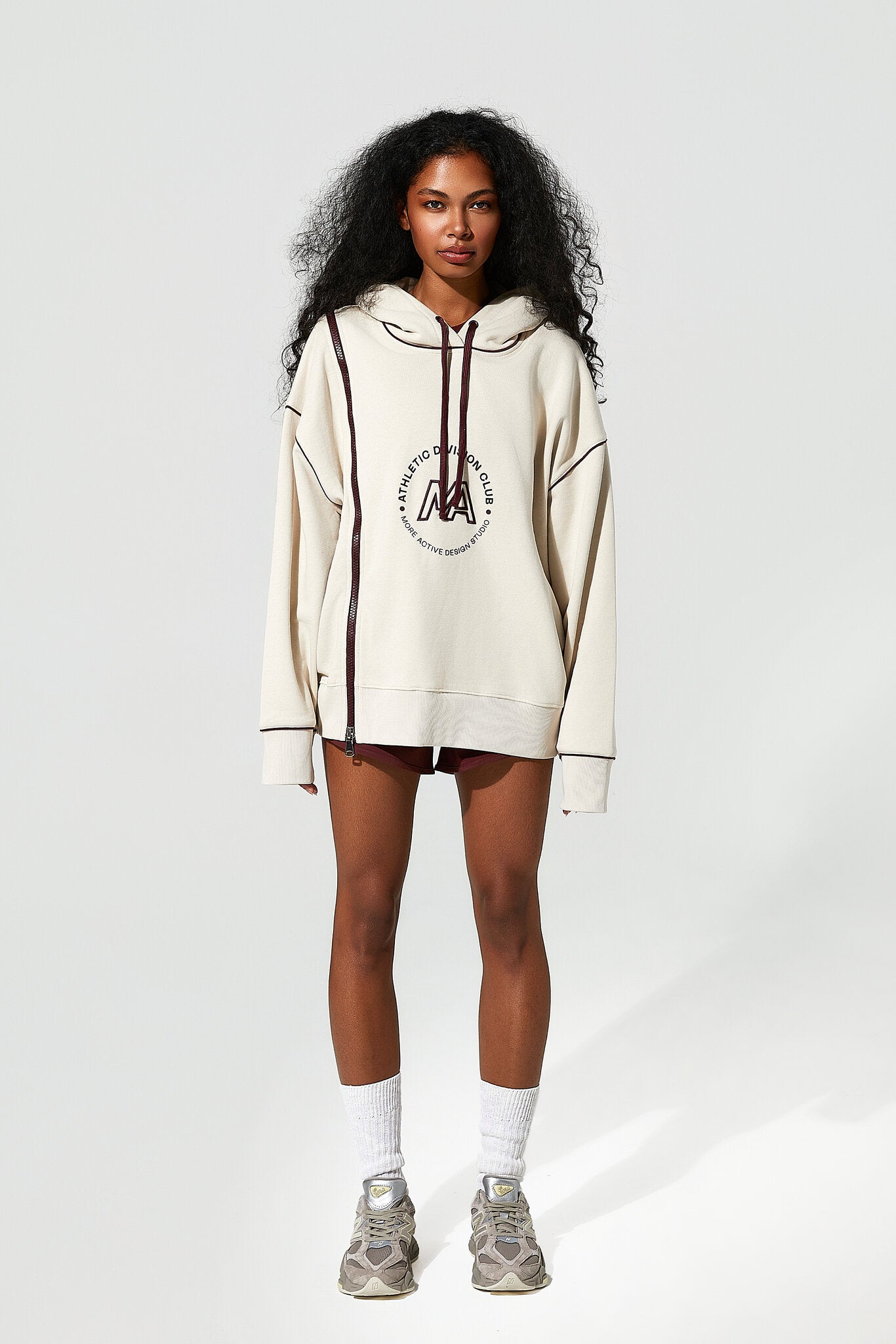 A model stands against a light gray studio background wearing an oversized cream hoodie with dark brown drawstrings and a circular chest graphic, paired with dark shorts, white socks, and gray sneakers.