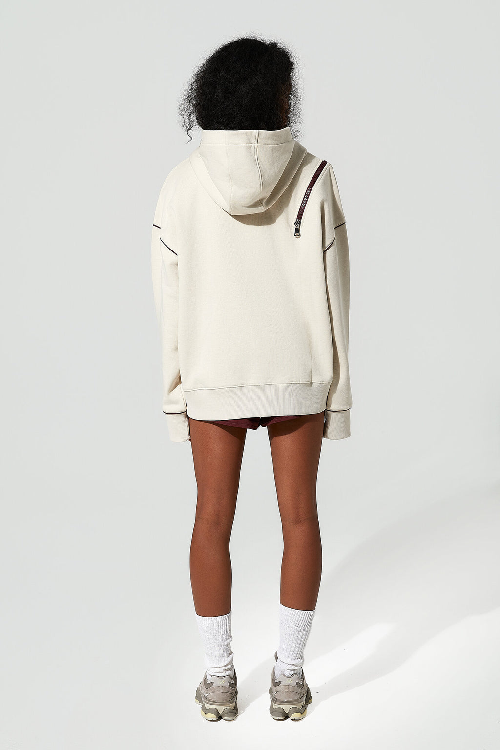 A model shown from the back wearing an oversized cream-colored hoodie with dark contrast stitching and a dark brown shoulder strap, paired with black shorts, white socks, and gray sneakers against a light gray background.