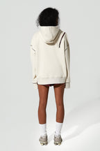 A model shown from the back wearing an oversized cream-colored hoodie with dark contrast stitching and a dark brown shoulder strap, paired with black shorts, white socks, and gray sneakers against a light gray background.