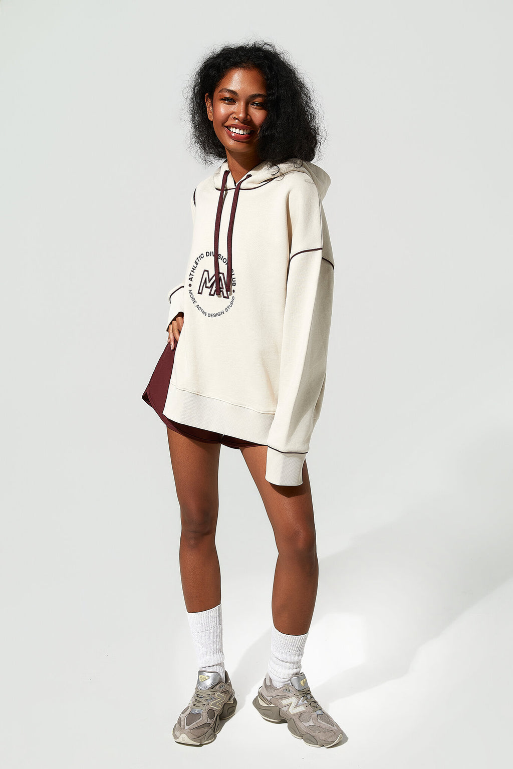 A woman modeling an oversized cream-colored hoodie with a dark red graphic print on the front, paired with a matching dark red mini skirt, white socks, and gray sneakers against a light gray studio background.