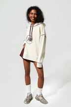 A woman modeling an oversized cream-colored hoodie with a dark red graphic print on the front, paired with a matching dark red mini skirt, white socks, and gray sneakers against a light gray studio background.