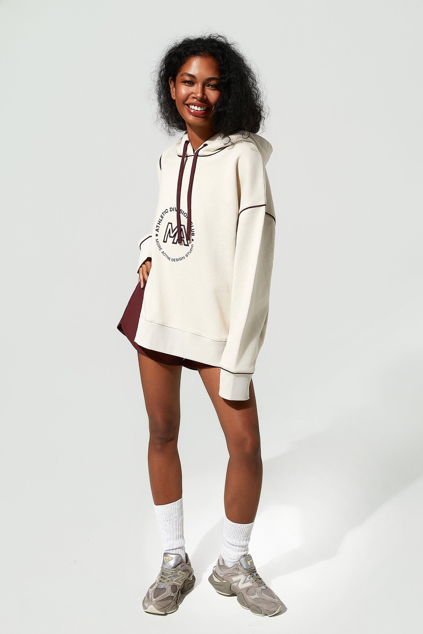A woman modeling an oversized cream-colored hoodie with a dark red graphic print on the front, paired with a matching dark red mini skirt, white socks, and gray sneakers against a light gray studio background.