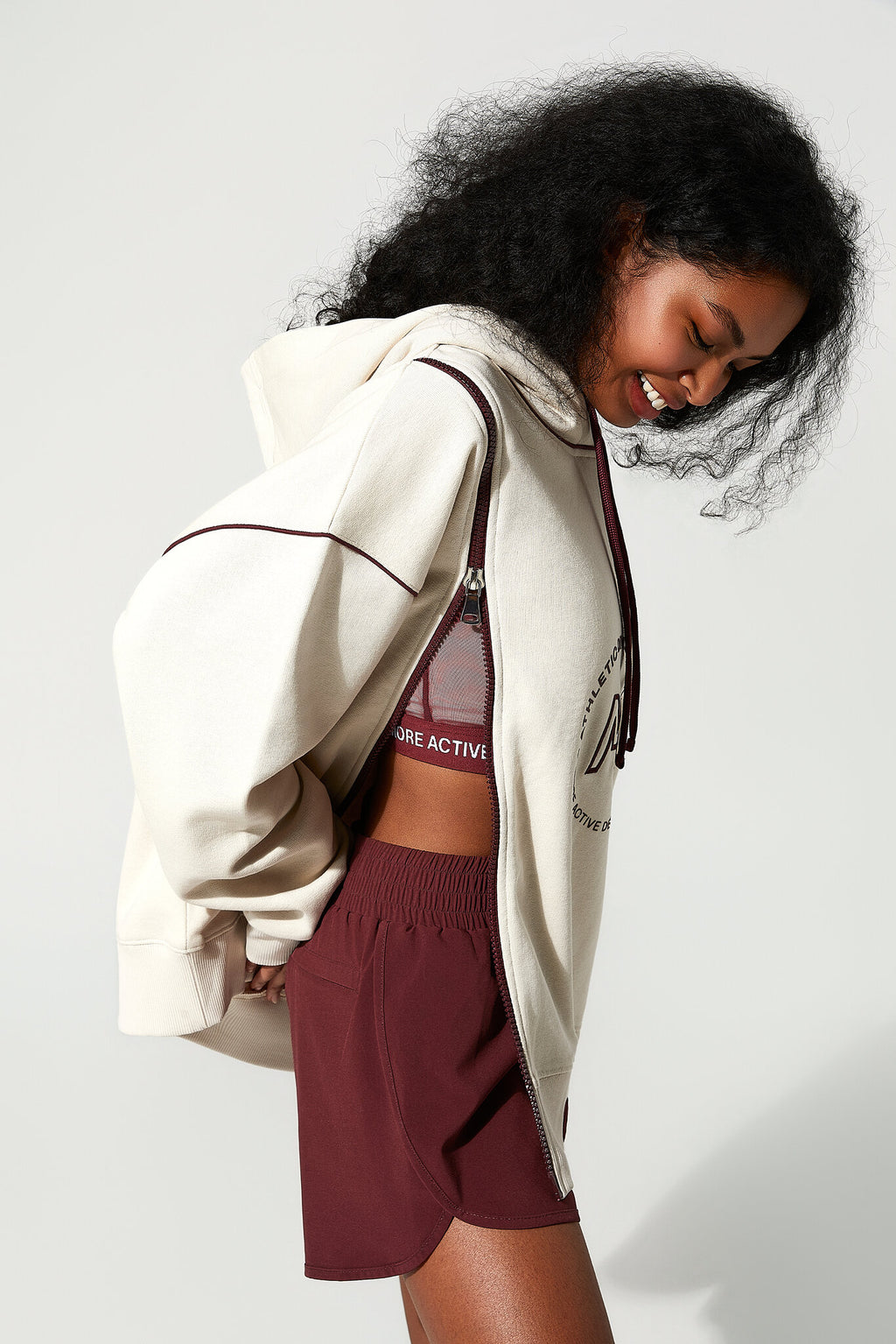 A woman wearing an oversized cream-colored zip-up hoodie with dark brown piping, burgundy athletic shorts, and a matching burgundy sports bra, posing against a light gray studio background.