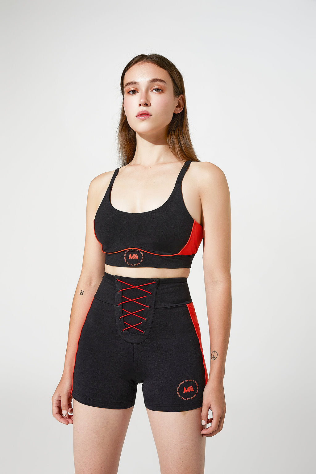 A woman modeling a matching black activewear set with red accents, including a sports bra and high-waisted shorts, against a light gray studio background.