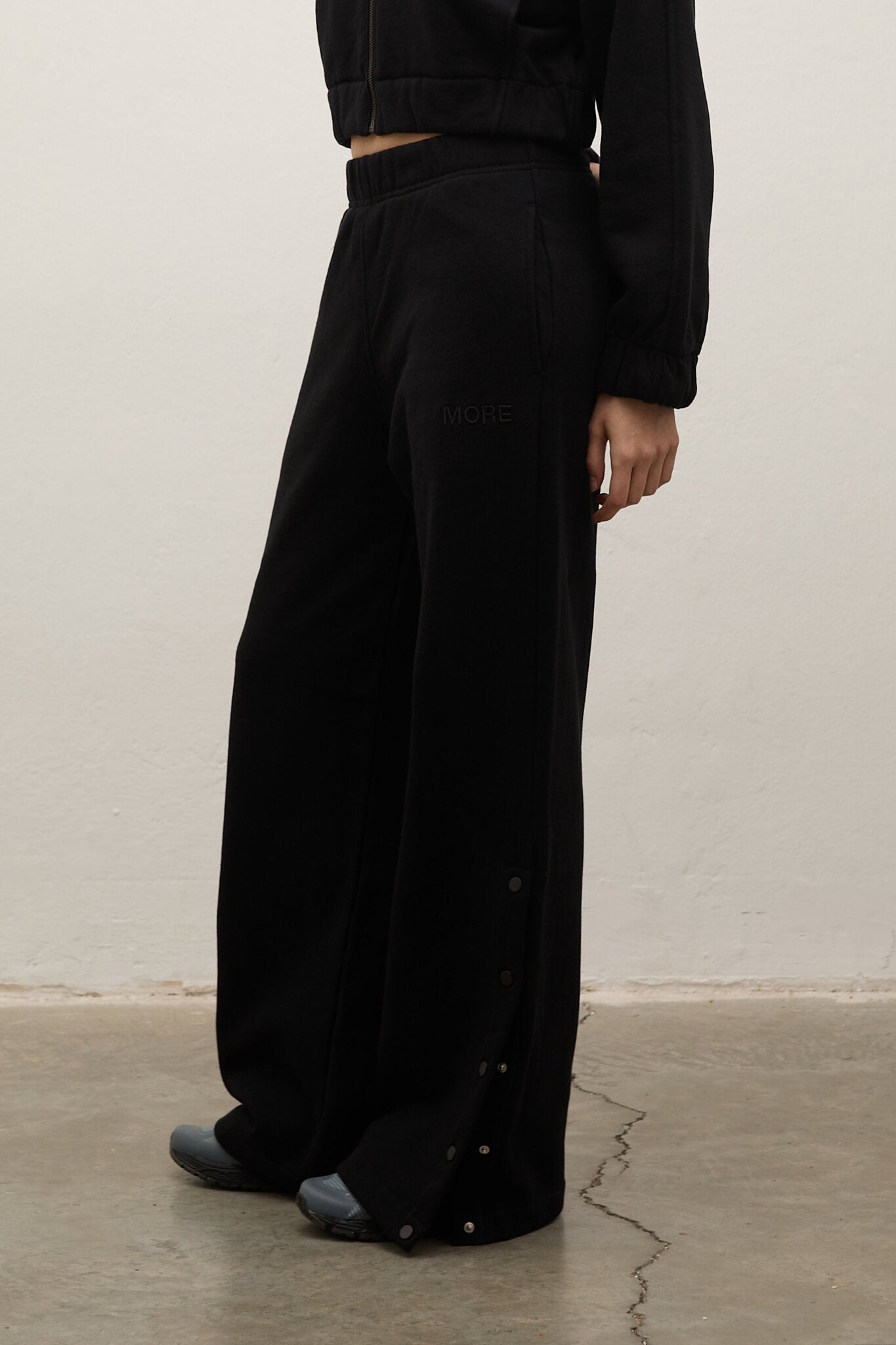A person wearing loose black wide-leg pants with visible side buttons, shown from the waist down against a plain off-white wall and concrete floor.