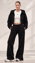 A model stands in a studio wearing a black cropped hooded jacket over a white cropped tank top, paired with black wide-leg pants and white sneakers.
