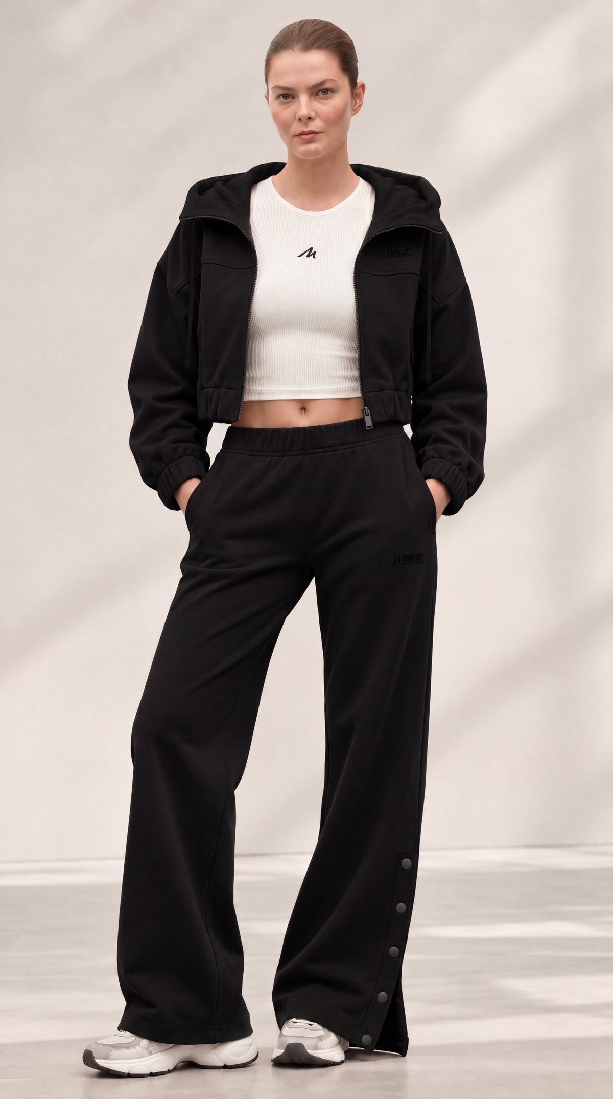 A model stands in a studio wearing a black cropped hooded jacket over a white cropped tank top, paired with black wide-leg pants and white sneakers.