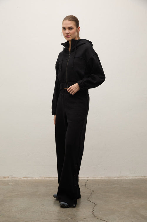 A model stands in a studio wearing a black zip-up hooded sweatshirt and matching black wide-leg pants against a plain off-white wall and concrete floor.