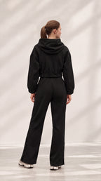 A woman viewed from behind wearing a black hooded jumpsuit with a drawstring waist and wide-leg pants, standing in a studio with a light gray background and soft shadow patterns.