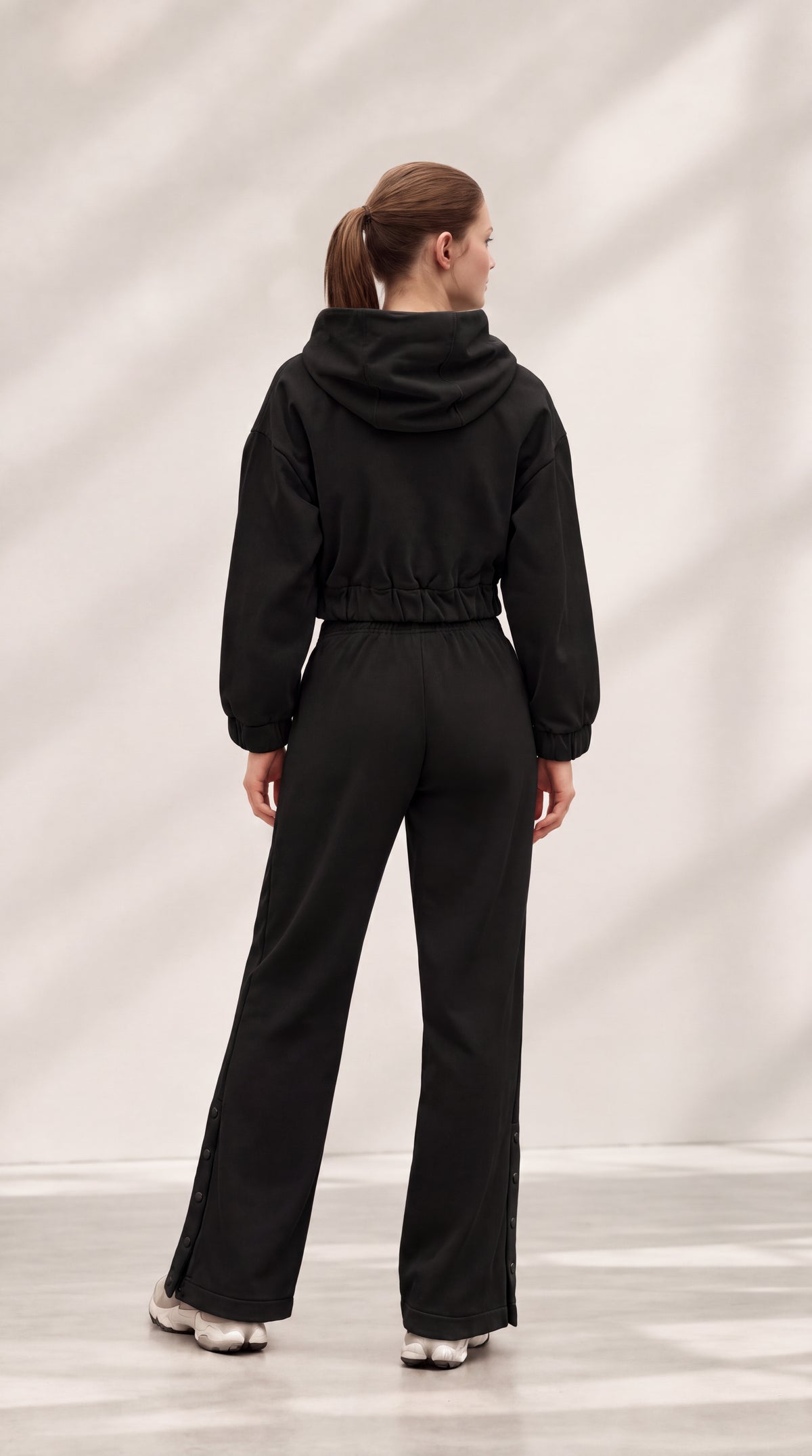 A woman viewed from behind wearing a black hooded jumpsuit with a drawstring waist and wide-leg pants, standing in a studio with a light gray background and soft shadow patterns.