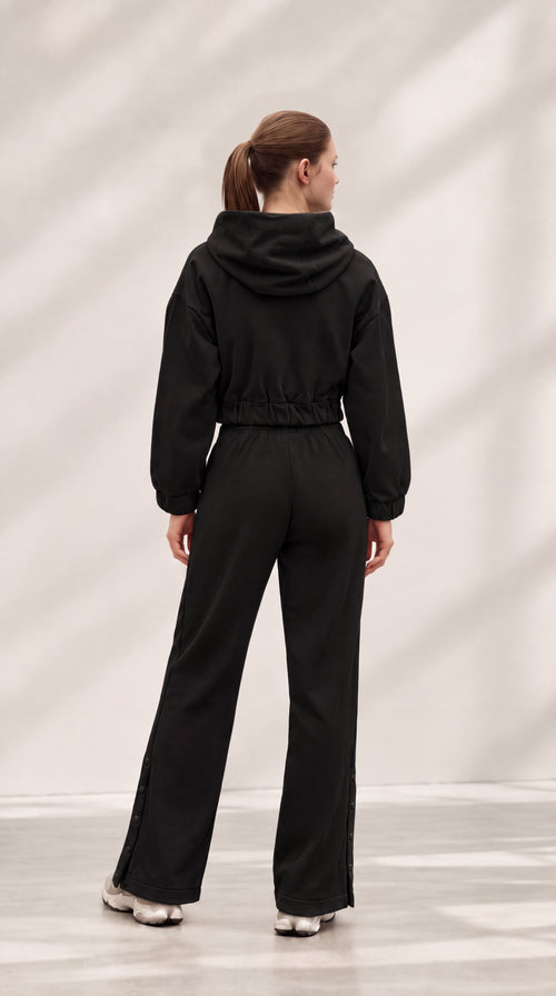 A woman viewed from behind wearing a black hooded jumpsuit with a drawstring waist and wide-leg pants, standing in a studio with a light gray background and soft shadow patterns.