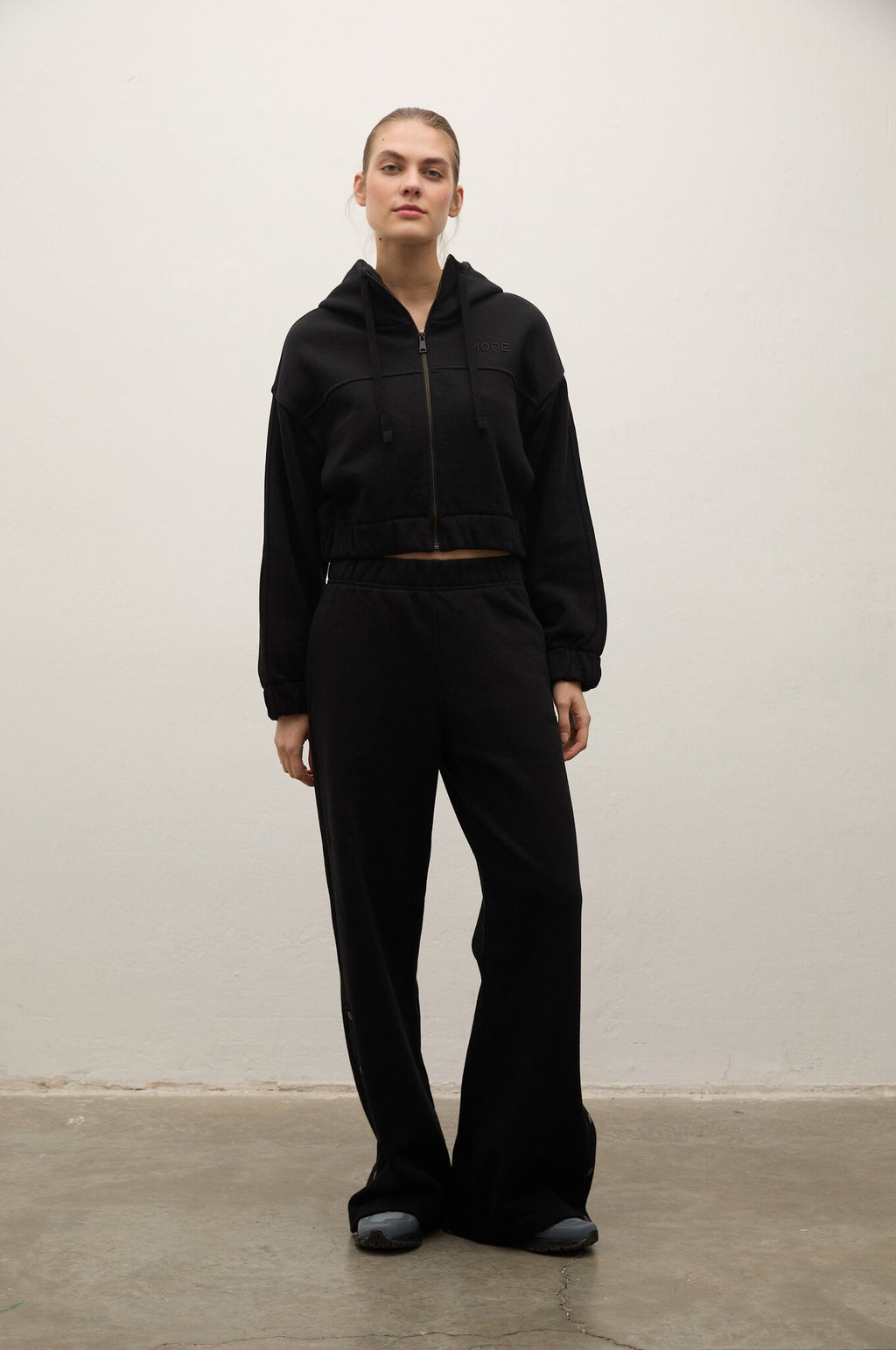 A model stands against a plain off-white wall wearing a black cropped zip-up hoodie and matching black wide-leg sweatpants.