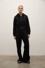 A model stands against a plain off-white wall wearing a black cropped zip-up hoodie and matching black wide-leg sweatpants.