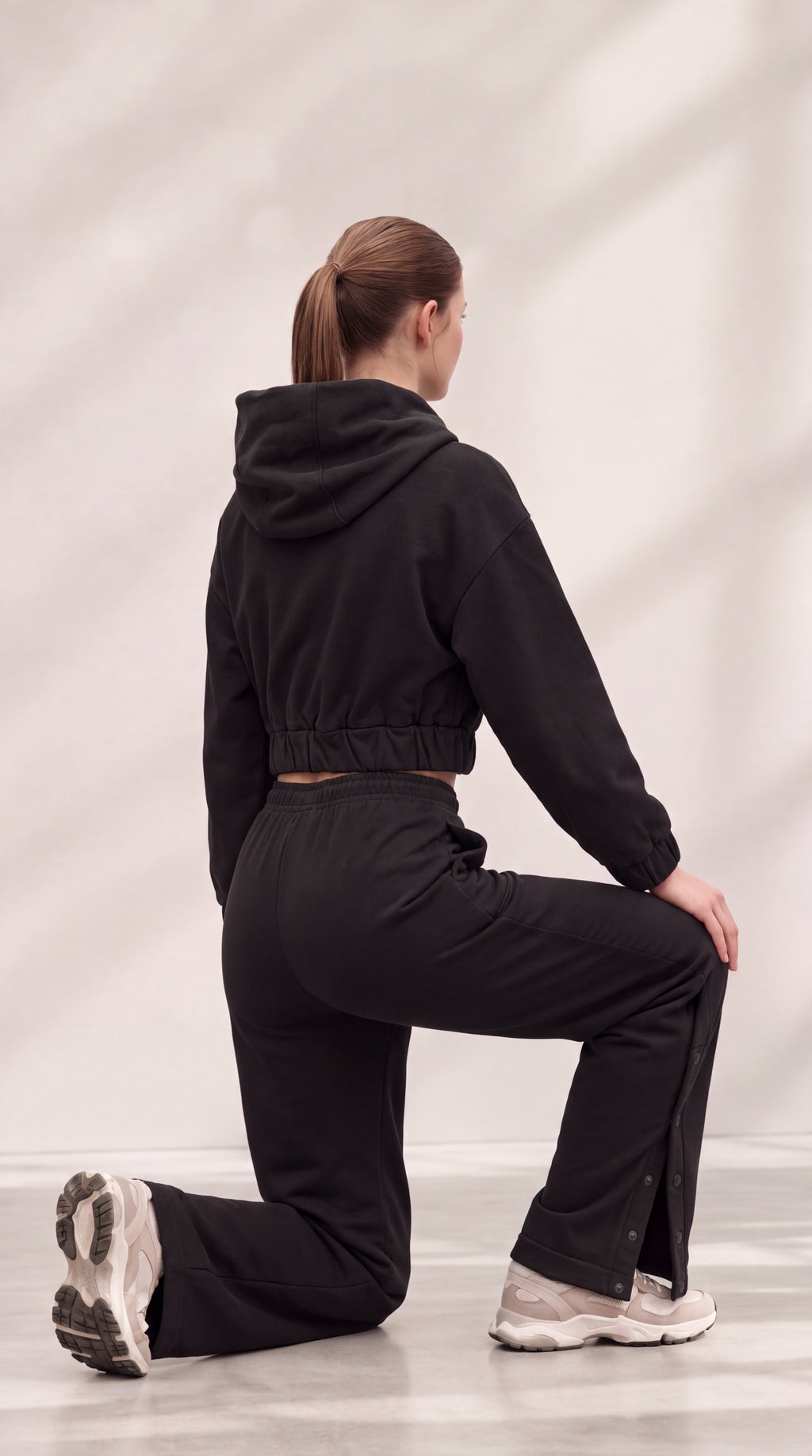 A woman kneeling in a black cropped hoodie and matching black pants, photographed from the back against a light gray studio backdrop with soft shadow overlays.