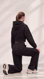 A woman kneeling in a black cropped hoodie and matching black pants, photographed from the back against a light gray studio backdrop with soft shadow overlays.