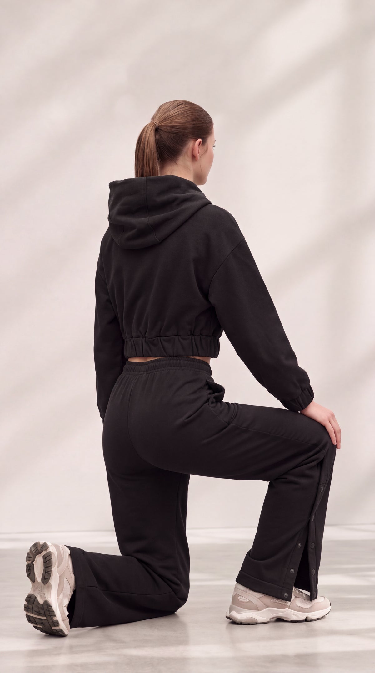 A woman kneeling in a black cropped hoodie and matching black pants, photographed from the back against a light gray studio backdrop with soft shadow overlays.