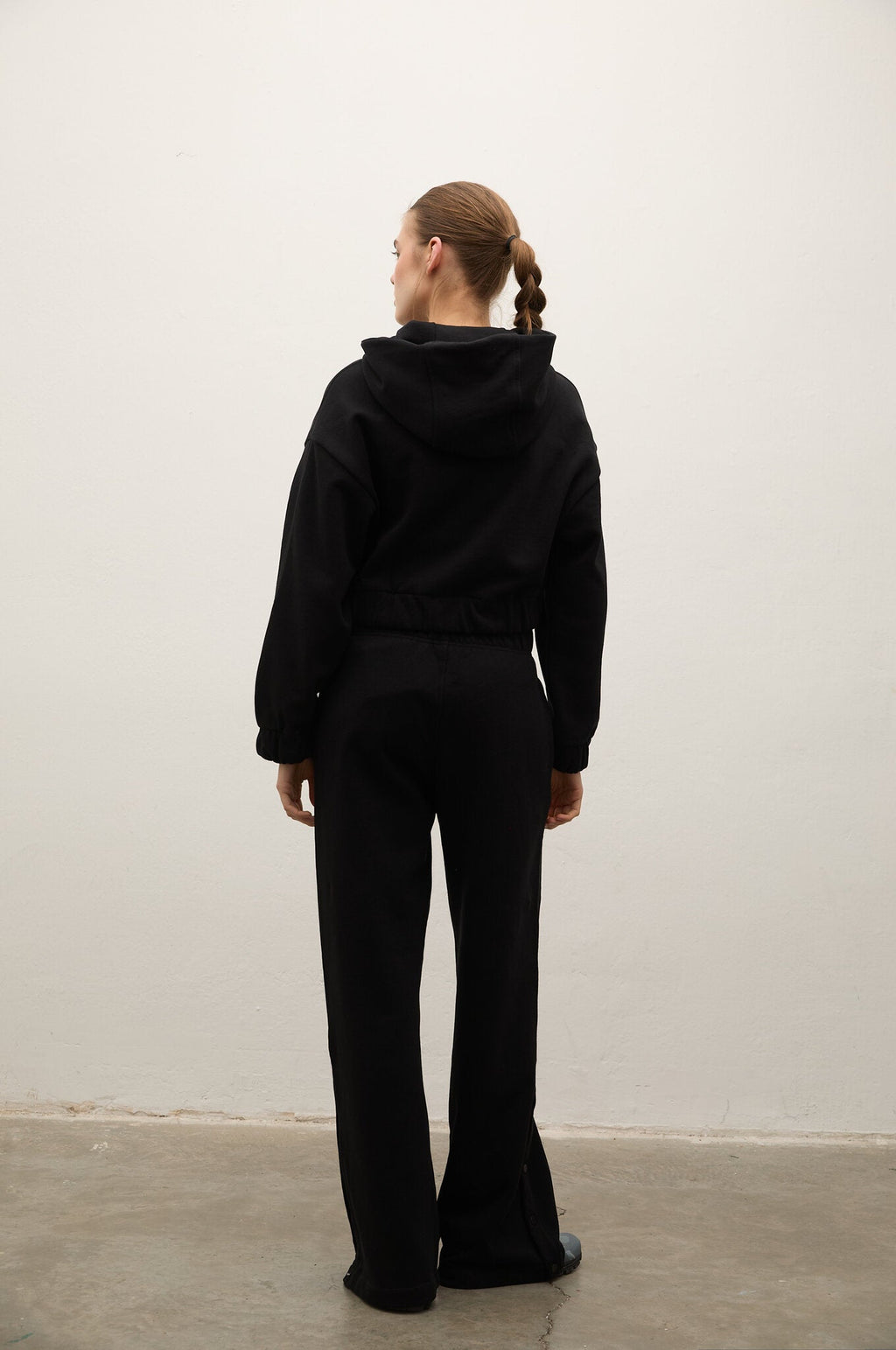 A woman viewed from behind wearing a black hooded sweatshirt and matching black wide-leg pants, standing against a plain off-white wall on a concrete floor.