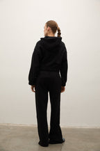 A woman viewed from behind wearing a black hooded sweatshirt and matching black wide-leg pants, standing against a plain off-white wall on a concrete floor.