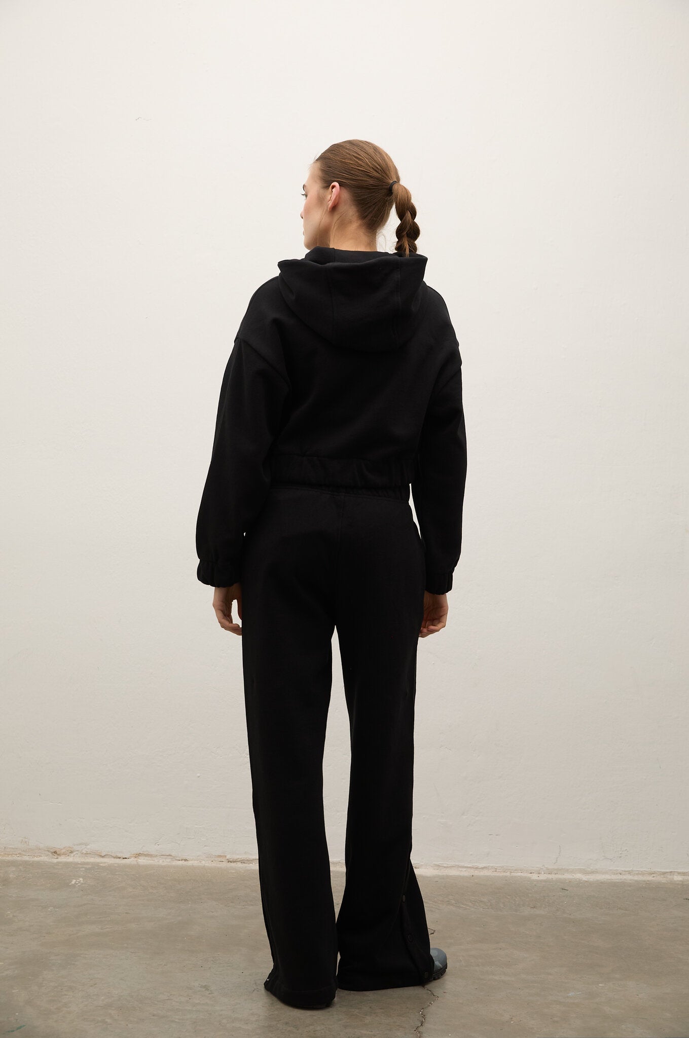 A woman viewed from behind wearing a black hooded sweatshirt and matching black wide-leg pants, standing against a plain off-white wall on a concrete floor.