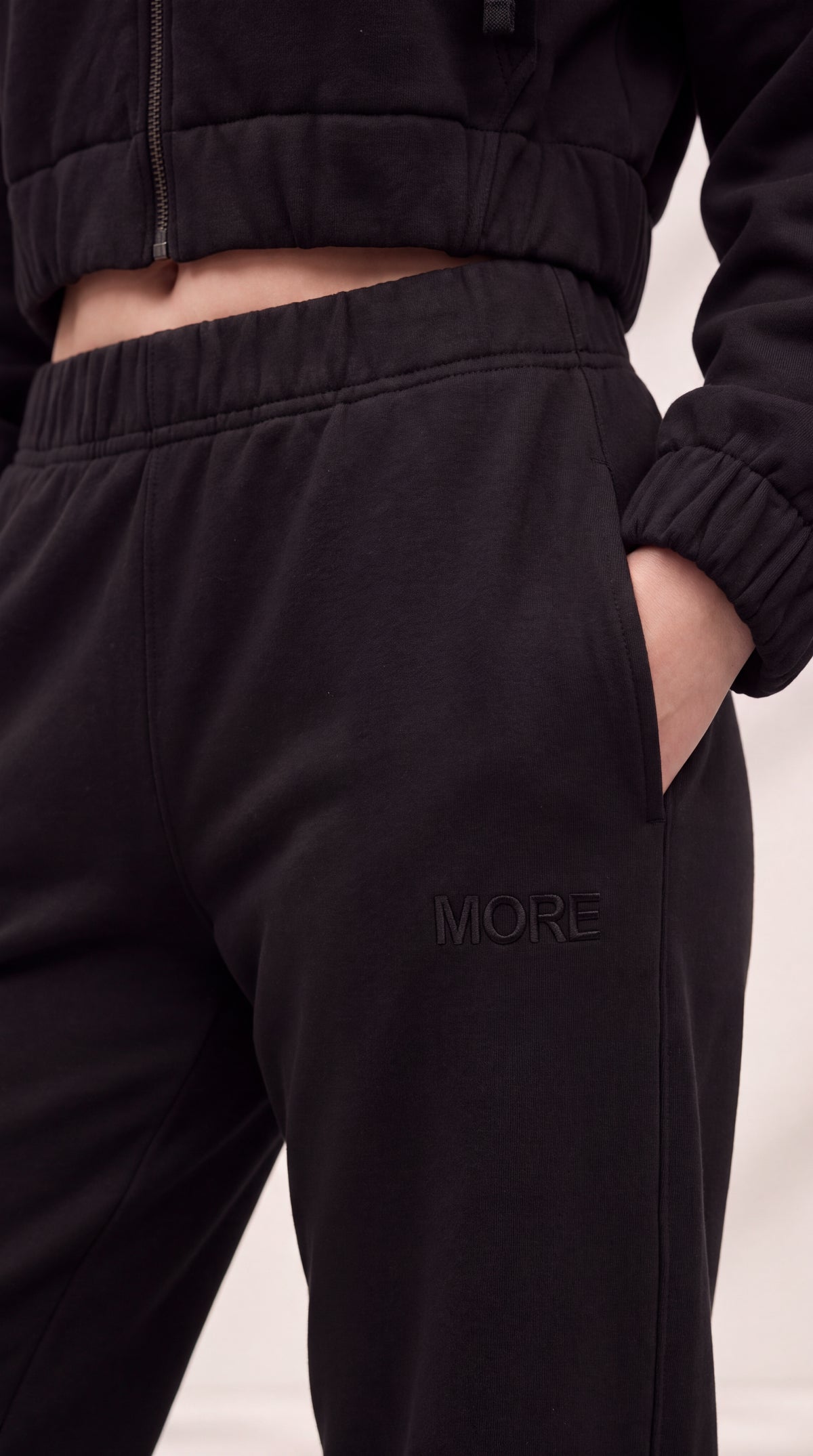 Close-up of a person wearing black jogger sweatpants with a side pocket and embossed 'MORE' branding, paired with a matching black cropped zip jacket.