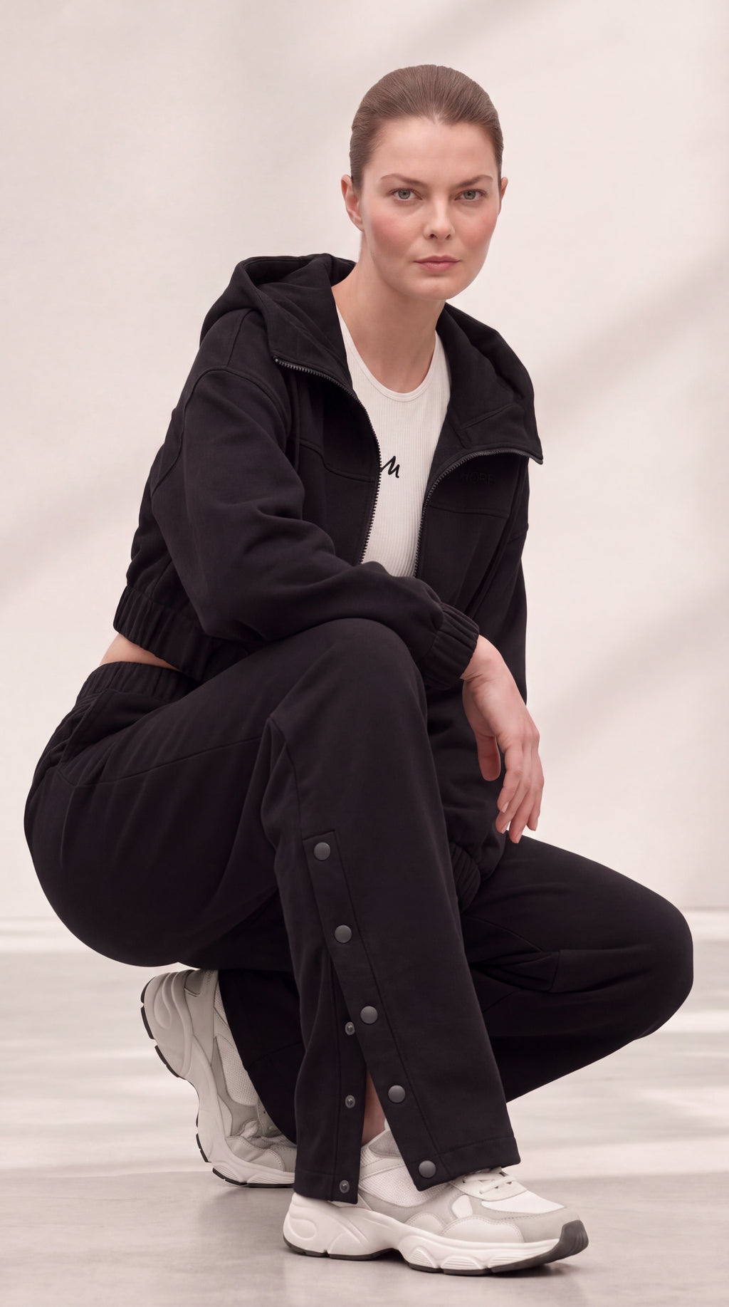 A model crouches in a studio wearing a black zip-up hoodie, black wide-leg pants with decorative metal snap details, a white t-shirt, and white sneakers.