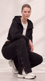 A model crouches in a studio wearing a black zip-up hoodie, black wide-leg pants with decorative metal snap details, a white t-shirt, and white sneakers.