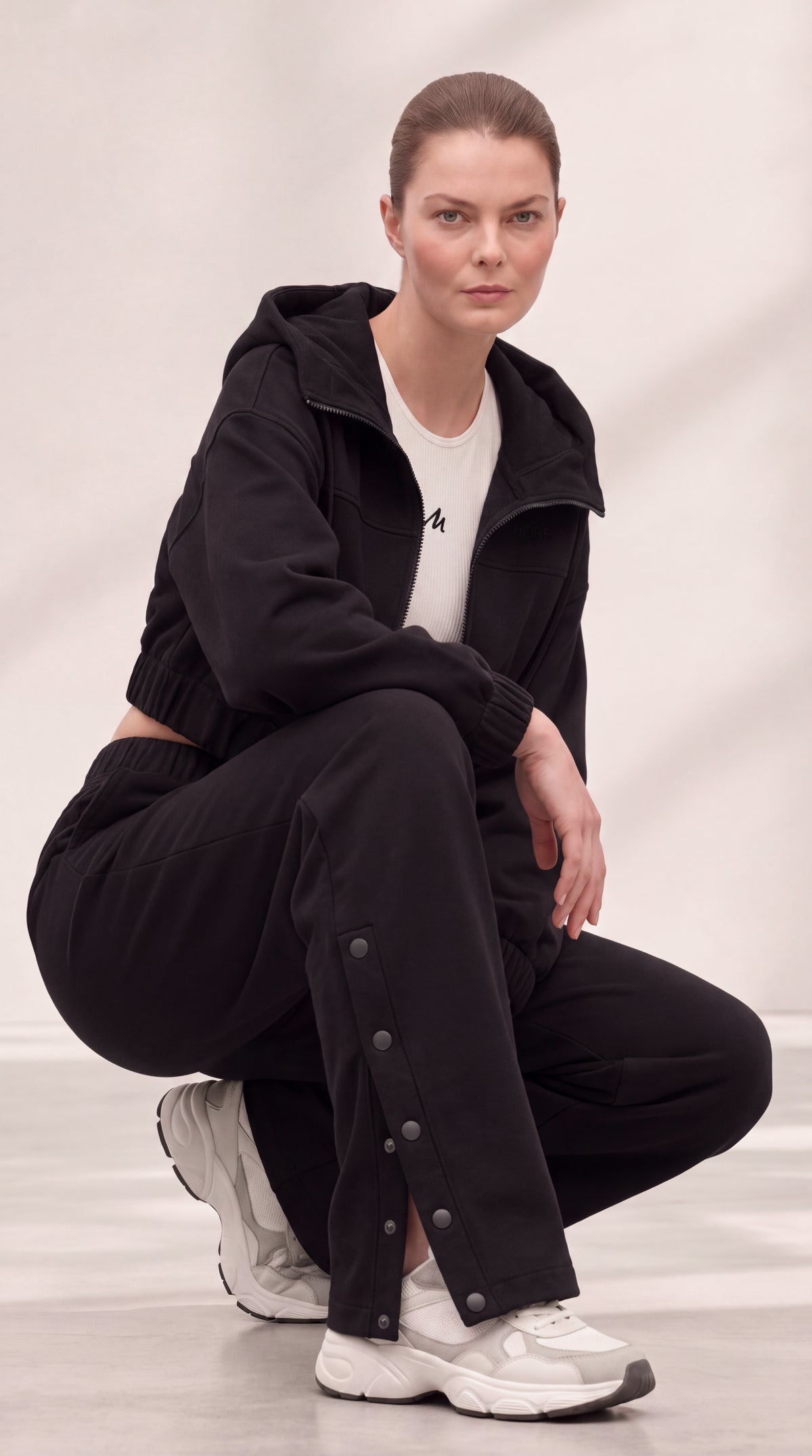 A model crouches in a studio wearing a black zip-up hoodie, black wide-leg pants with decorative metal snap details, a white t-shirt, and white sneakers.