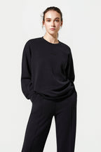 A woman modeling a black crewneck sweatshirt and matching black wide-leg trousers against a plain light gray studio background.