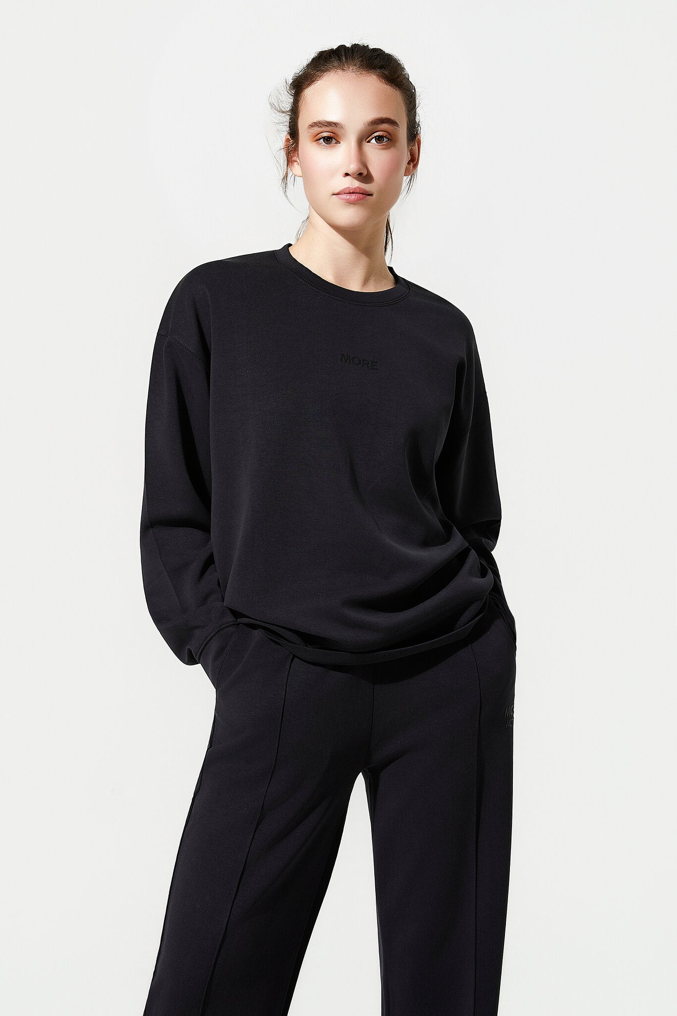 A woman modeling a black crewneck sweatshirt and matching black wide-leg trousers against a plain light gray studio background.
