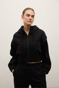 A woman modeling a black cropped zip-up hoodie with drawstrings and a subtle tonal logo on the chest, paired with black sweatpants, against a plain light gray studio background.