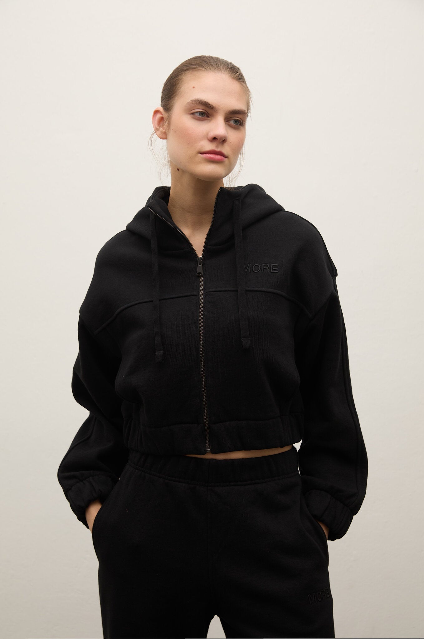 A woman modeling a black cropped zip-up hoodie with drawstrings and a subtle tonal logo on the chest, paired with black sweatpants, against a plain light gray studio background.